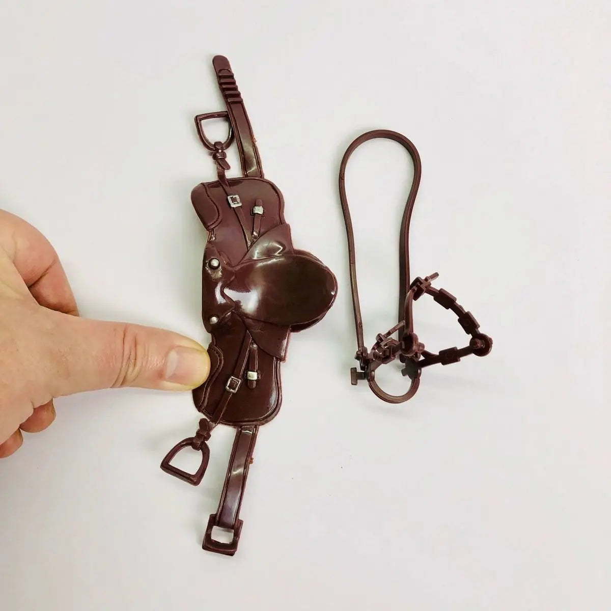 A brown leather dollhouse horse saddle and riding prop accessory being held by a hand.