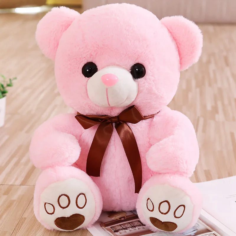 A pink plush teddy bear with black eyes and a brown bow sitting on a wooden surface.