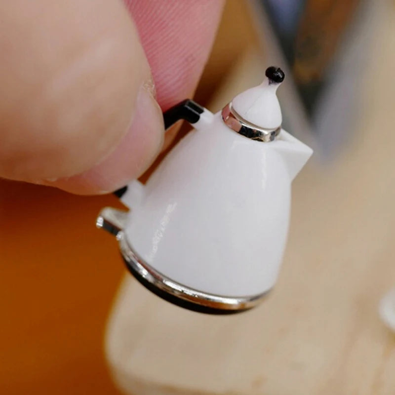 A hand holds a miniature silver alloy kettle with a white body and black spout, used for dollhouse kitchen decor.