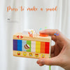 A hand holds a wooden camera teether toy with colorful stripes and smiling faces, designed to make a sound.