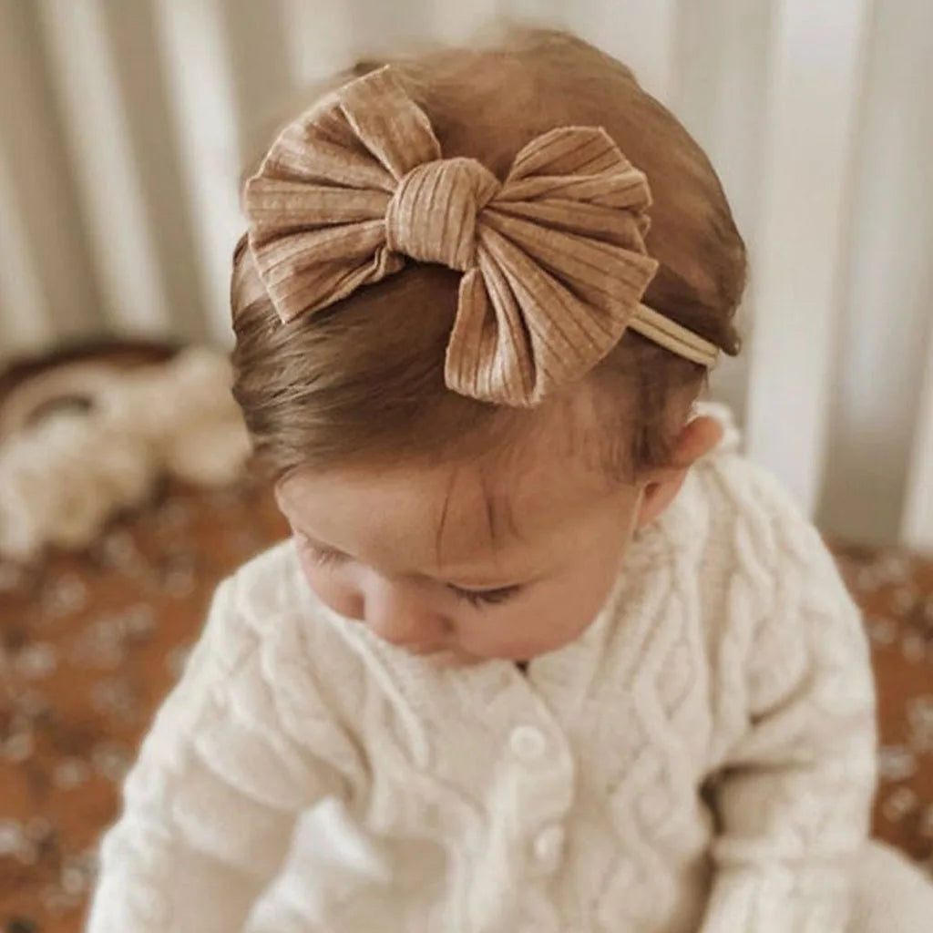 A baby girl wearing a beige bow headband and a white knitted sweater.