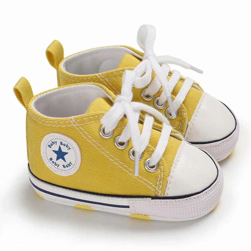 A pair of yellow canvas baby shoes with white laces and soles are shown against a white background.