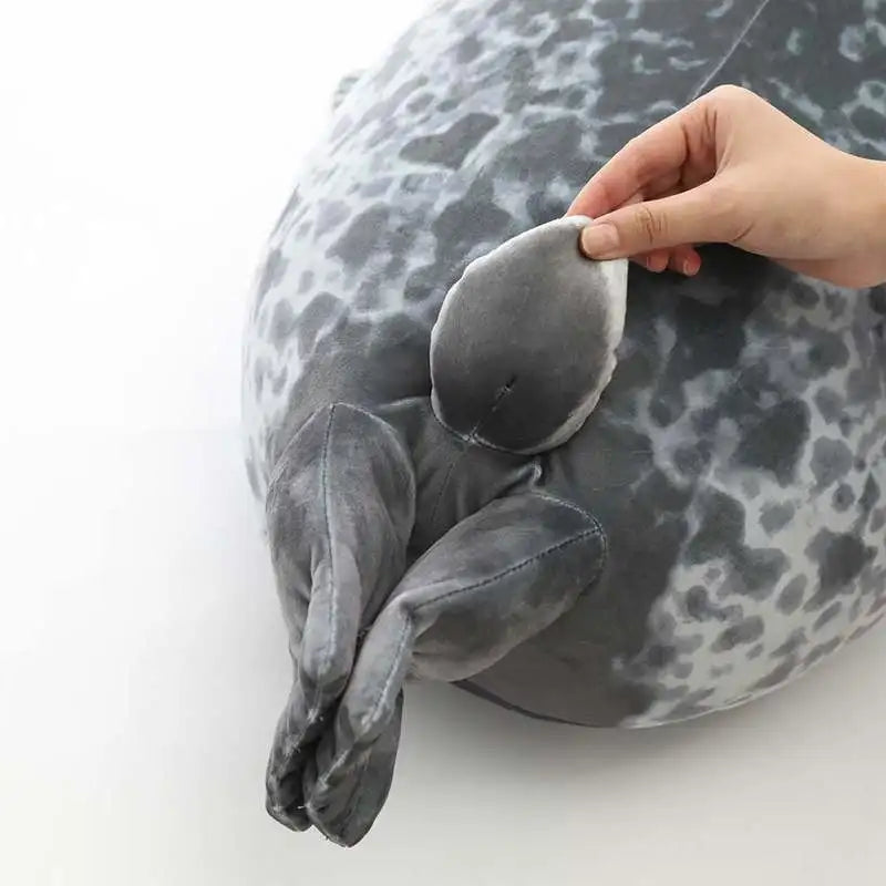 A hand is gently pulling a gray and white spotted plush seal pillow toy.