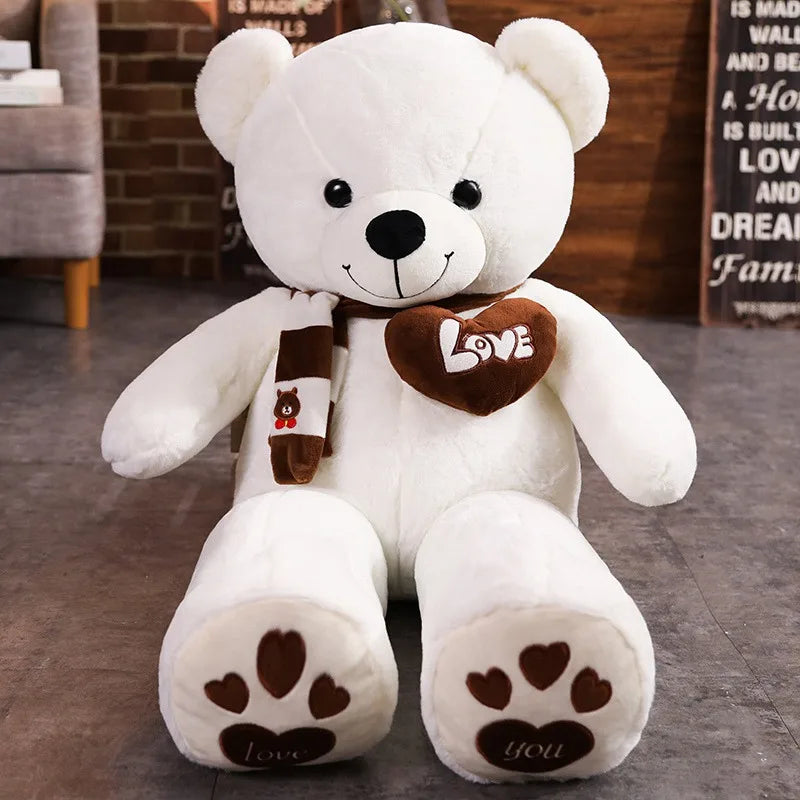 A white plush teddy bear wearing a brown heart-shaped accessory and knit scarf sitting on a gray floor.