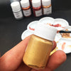A hand holds a bottle of metallic gold acrylic paint for crafts, with several small bottles in the background.