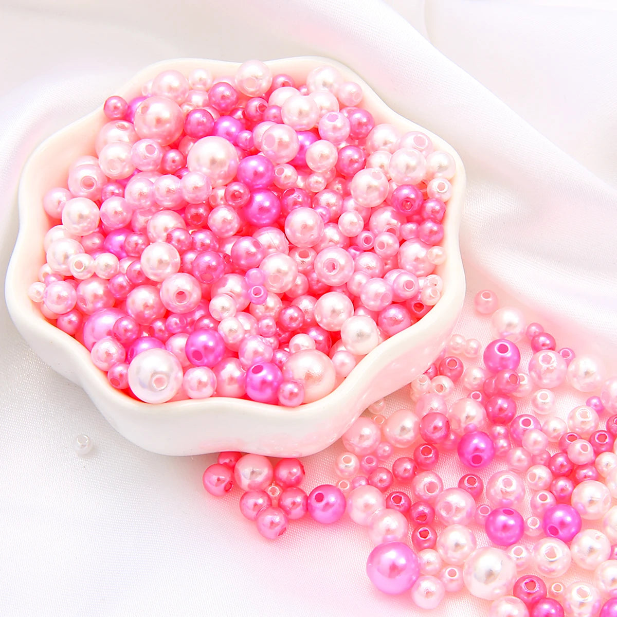 A pile of assorted pink and magenta pearl beads in a white dish and scattered on a white surface.