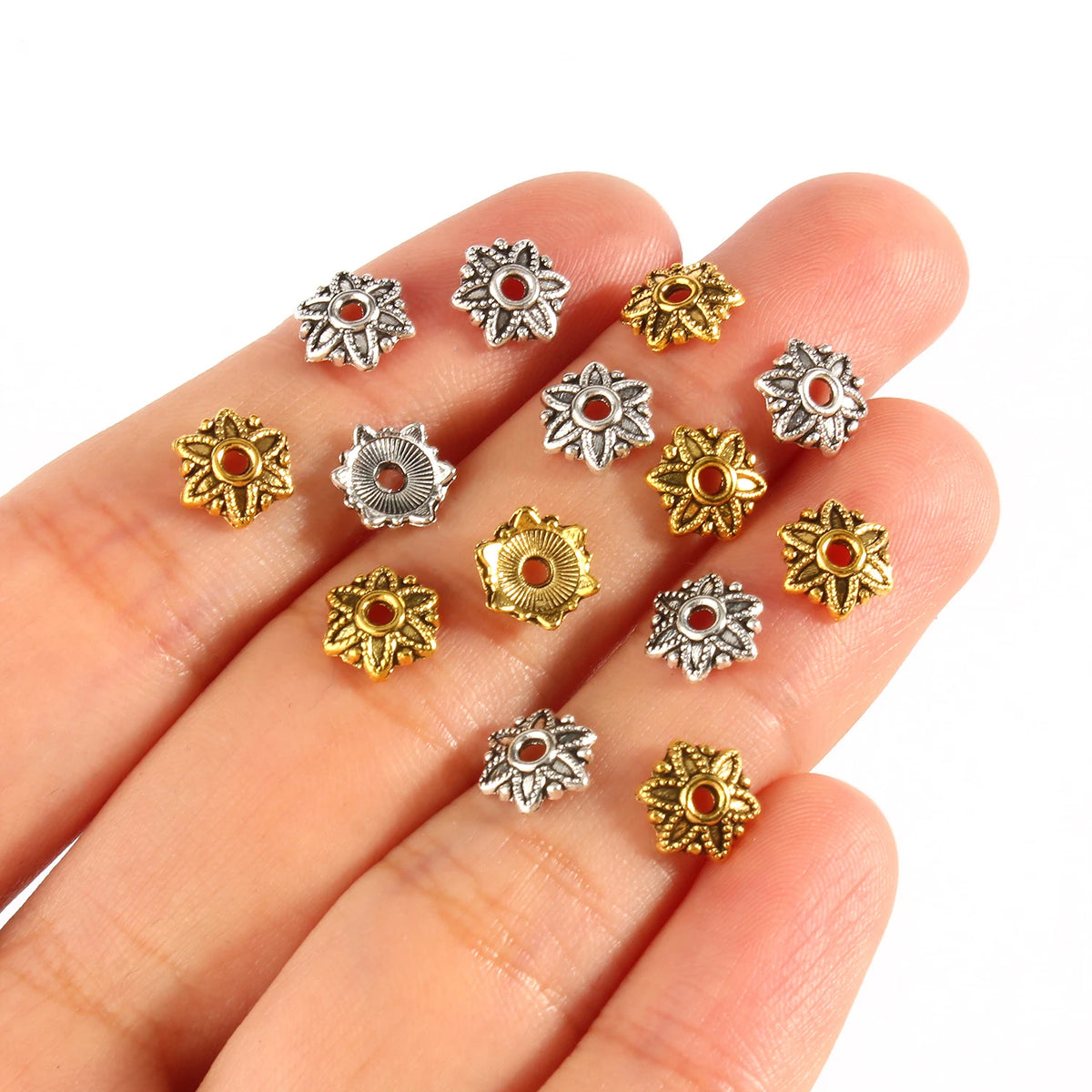 A hand holds several small, silver and gold Tibetan style hollow flower bead caps for jewelry making.