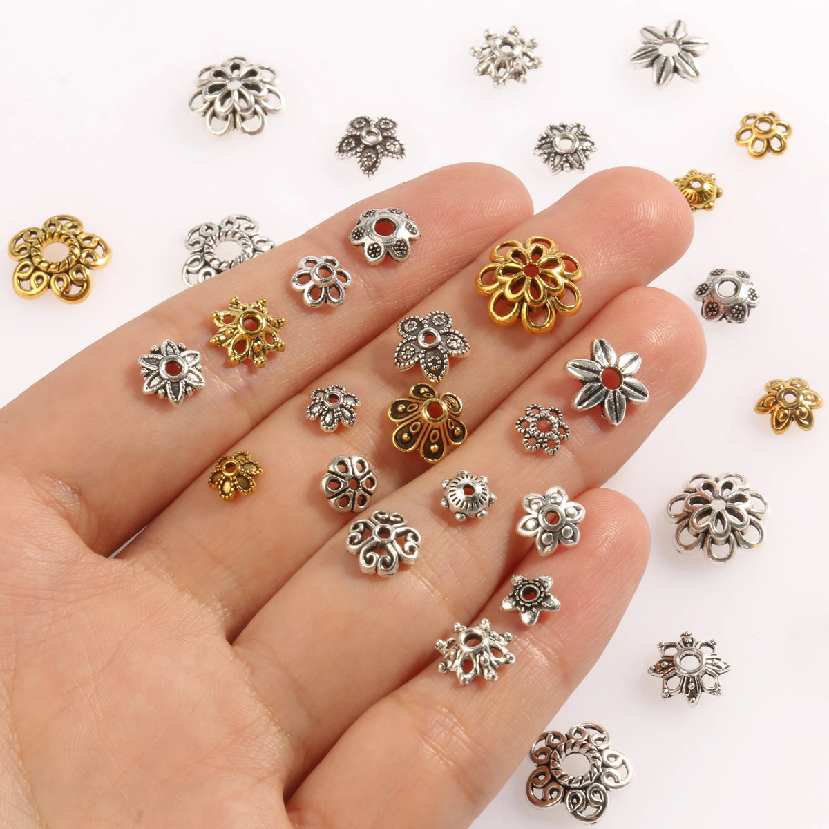 A hand displays various silver and gold Tibetan style hollow flower bead caps for jewelry making.