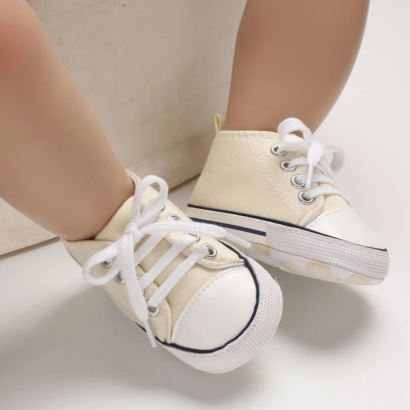 A pair of beige canvas first walkers for infants with white laces and a speckled sole.
