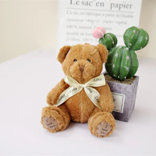 A soft brown teddy bear plush toy with a light yellow ribbon and a cactus decoration sitting in a small box.