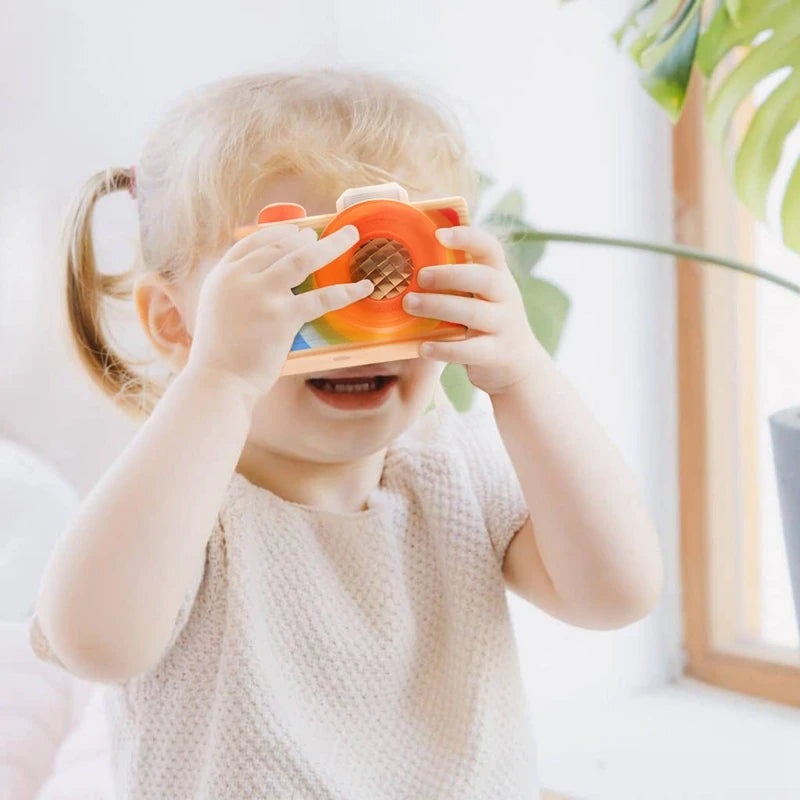A young girl is holding and looking through an orange wooden camera teether toy.