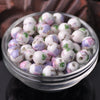 A clear glass bowl filled with small ceramic beads in shades of white, pink, and purple flowers for jewelry making