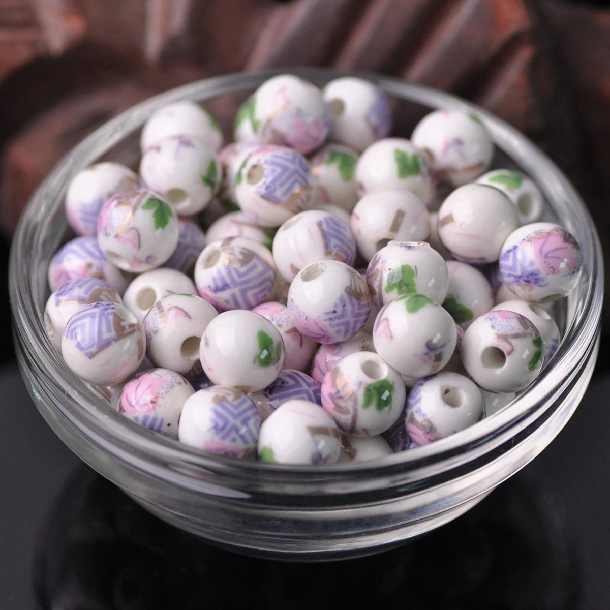 A clear glass bowl filled with small ceramic beads in shades of white, pink, and purple flowers for jewelry making