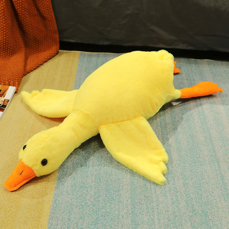 A yellow oversized kawaii goose plush pillow with orange beak and feet lying on a light blue surface.