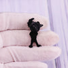 A black enamel pin shaped like a cartoon character dog is held between white gloves.