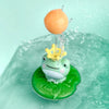 An orange sphere is being sprayed by a yellow crown from a green frog bath toy floating in water.