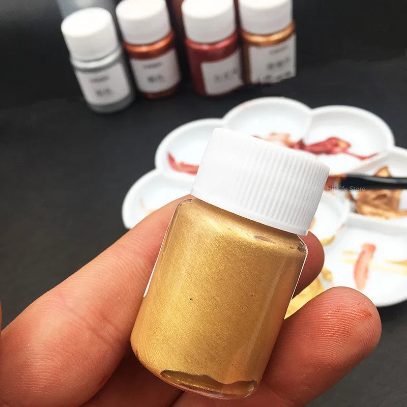A hand holds a bottle of metallic gold acrylic paint for crafts, with several small bottles in the background.