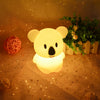 A glowing white koala LED night light sits on a textured surface surrounded by green foliage and scattered gold stars.