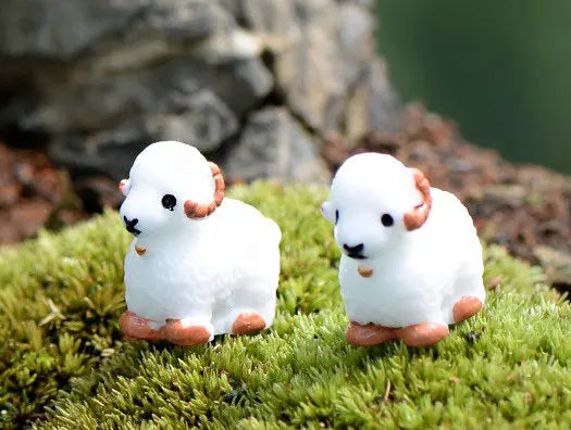 Two white miniature lamb figurines with pink accents resting on green moss.