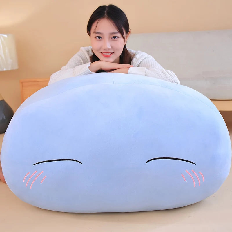 A light blue, oval-shaped plush toy pillow resembling a sleeping anime slime character with closed eyes and pink eyelashes.
