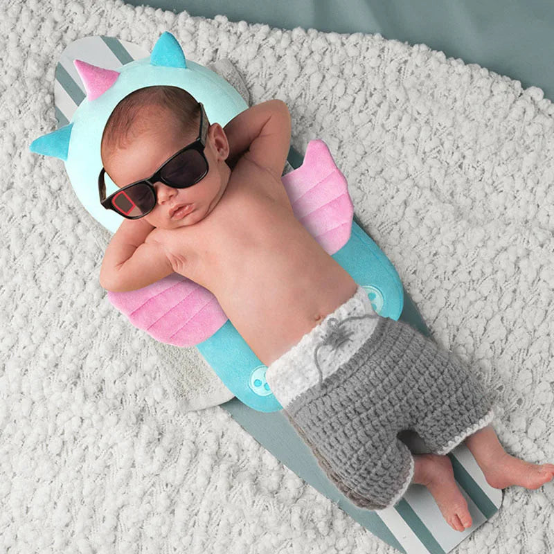A baby is lying on a soft gray blanket, wearing sunglasses and a light blue head protector pillow with pink straps.