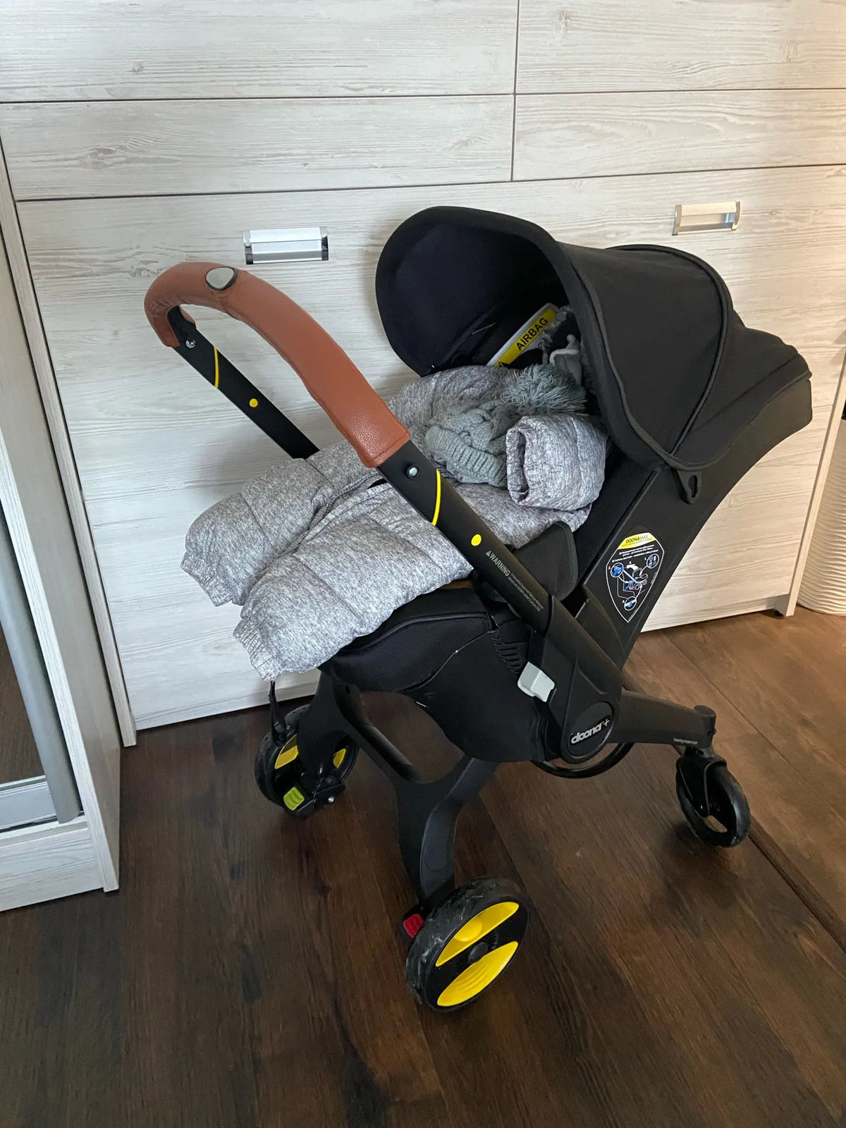 A black stroller accessory kit featuring a mosquito net, rain cover, and other items resting on a wooden floor.