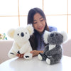 A young woman is holding two plush koala toys, one white and one gray, on a white table.