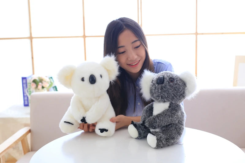 A young woman is holding two plush koala toys, one white and one gray, on a white table.