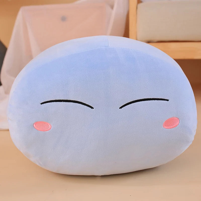 A light blue, round plush toy pillow shaped like a character with closed eyes and pink cheeks.