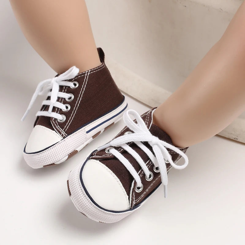 A pair of brown canvas first walkers for infants with white laces and soles, shown on a person's foot.