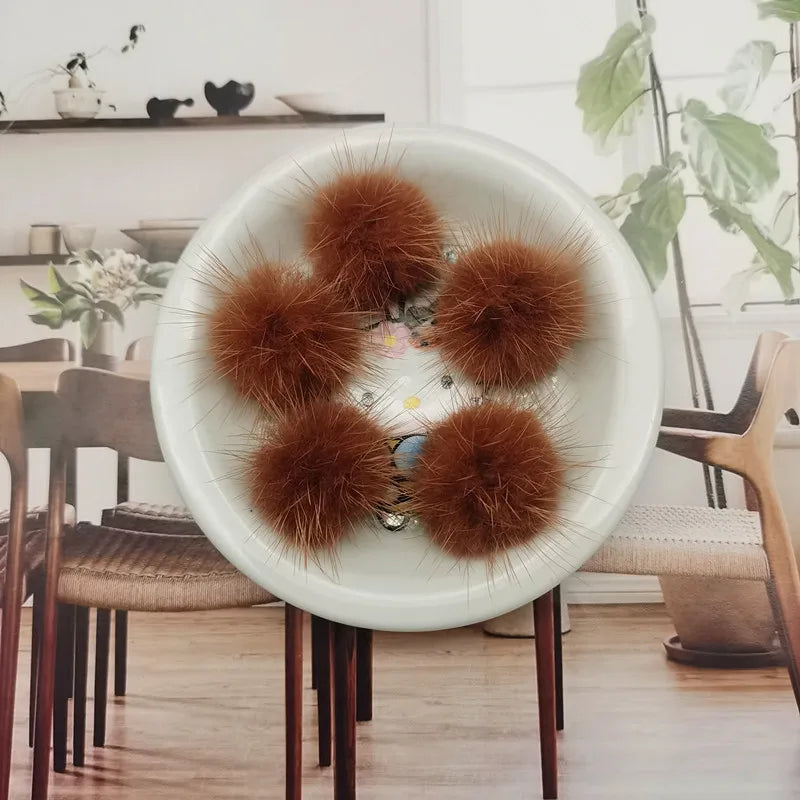 A white bowl filled with several fluffy, reddish-brown mink fur pom poms for crafts and accessories.