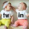 Two babies wearing white letter print bodysuits, 'tw' and 'in', sitting together.