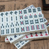 A stack of white Mahjong tiles with various symbols and red markings is displayed on a textured paper background.