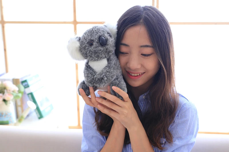 A young woman is holding a gray koala plush toy against her chest in a bright indoor setting.