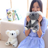 A young woman is holding a gray and white koala plush toy in front of her, with another white plush toy on the sofa beside he