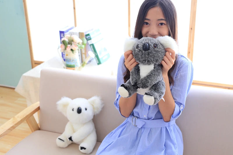 A young woman is holding a gray and white koala plush toy in front of her, with another white plush toy on the sofa beside he