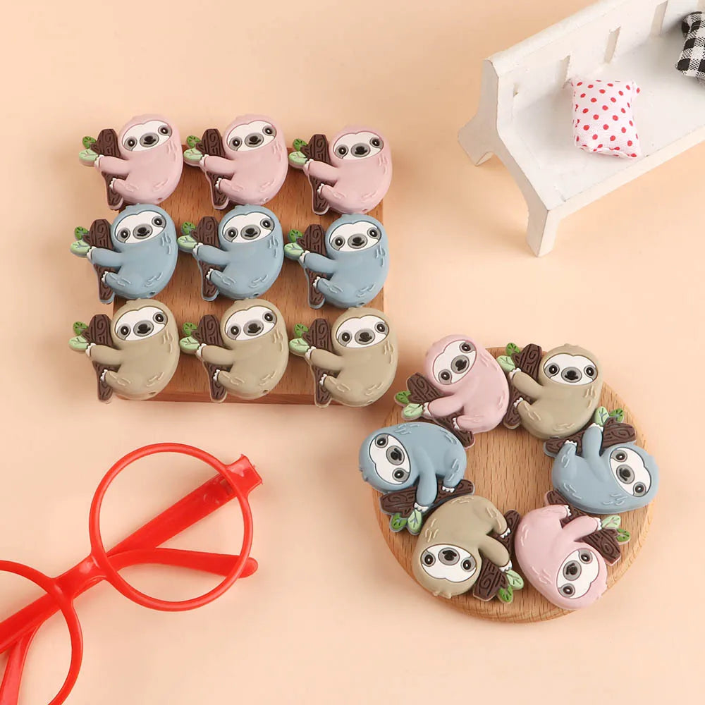 A collection of adorable sloth silicone beads in various shades of gray and pink arranged on a wooden board.
