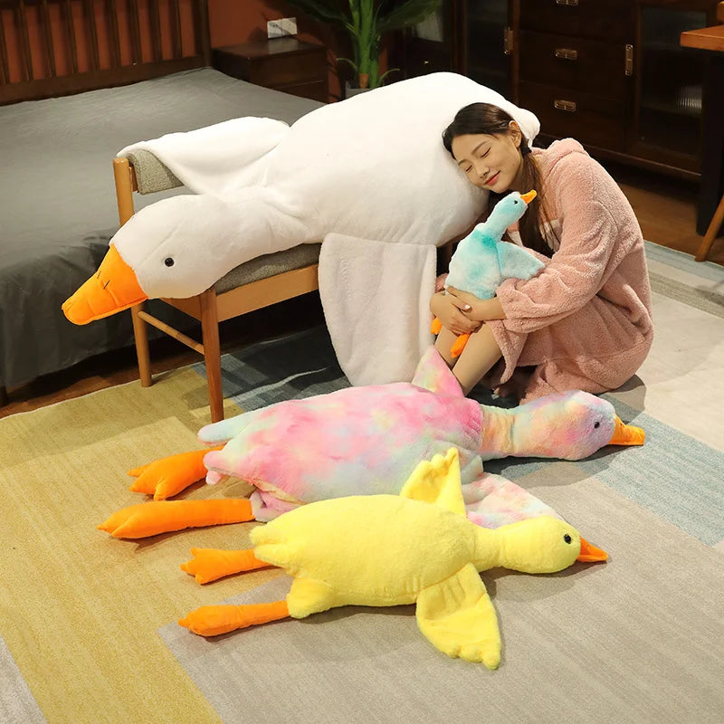 A woman is relaxing with an oversized white goose plush pillow and several colorful yellow and pink goose toys on a light rug