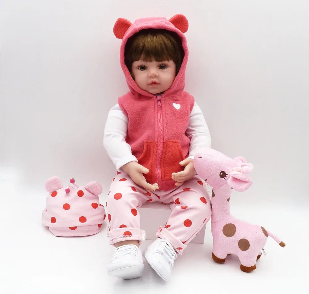 A baby doll wearing a pink hooded jacket and matching pants sits next to a pink stuffed giraffe.