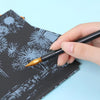A hand holds a paintbrush applying gold paint to a black scratch art design on paper.