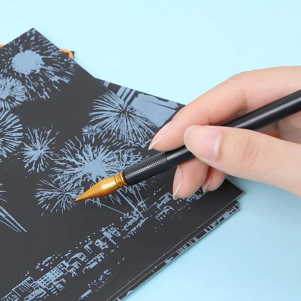 A hand holds a paintbrush applying gold paint to a black scratch art design on paper.