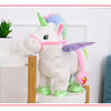 A white unicorn plush toy with pink accents and a green horn standing next to a potted plant.