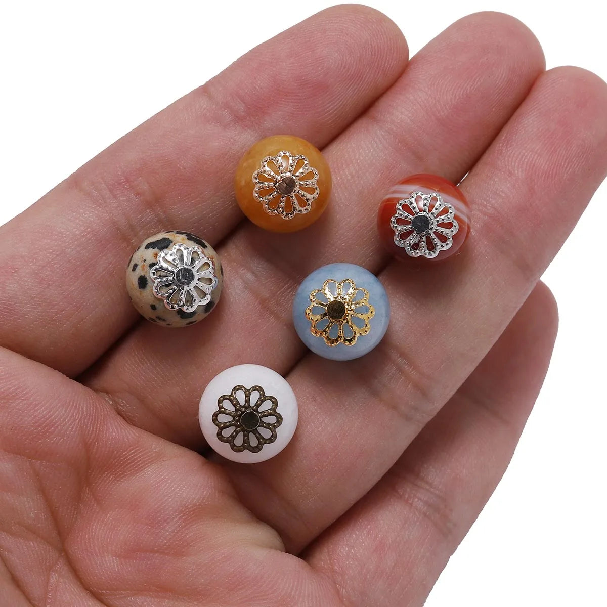 A hand holds several gold-plated flower petal bead caps in various shades of orange, white, and blue.