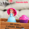 A pink cupcake doll with a blue dress is displayed in a wooden box next to a purple cupcake liner.