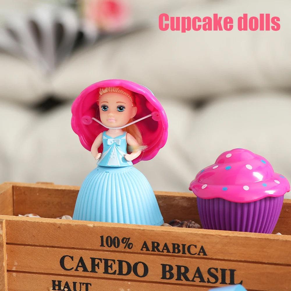 A pink cupcake doll with a blue dress is displayed in a wooden box next to a purple cupcake liner.