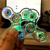 A hand holds a glow-in-the-dark LED fidget spinner with multiple light-up kinetic components.