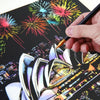 A hand holds a paintbrush applying paint to a scratch art piece featuring a cityscape and fireworks.