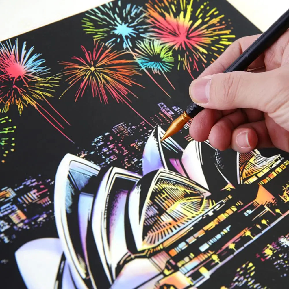 A hand holds a paintbrush applying paint to a scratch art piece featuring a cityscape and fireworks.