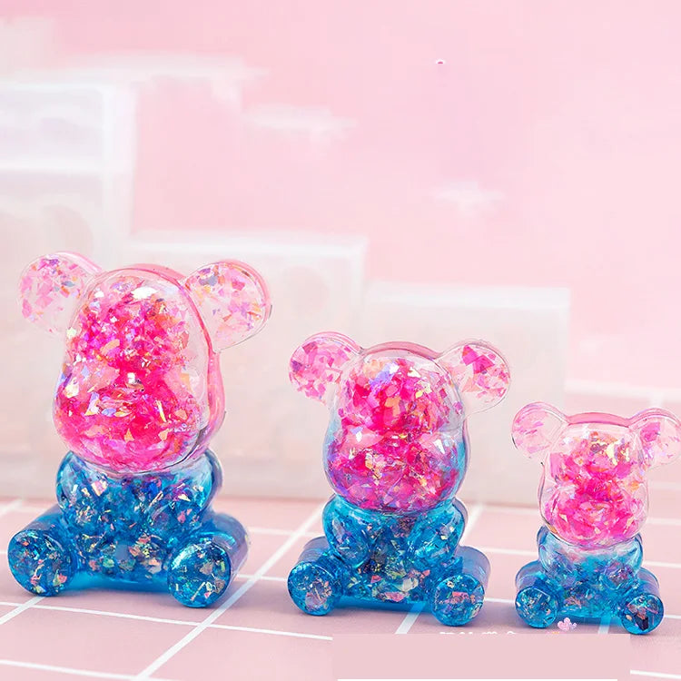 Three small, bear-shaped silicone molds in pink and blue glitter are displayed on a light pink surface.