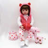 A baby-like reborn doll wearing a pink hooded outfit and pink pants, sitting on a white background.